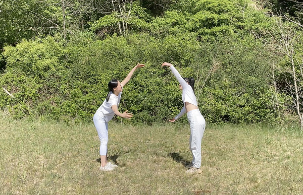 In the forest, two people are opening arms and fronting each other with distance. They are wearing white clothes and have black hair.